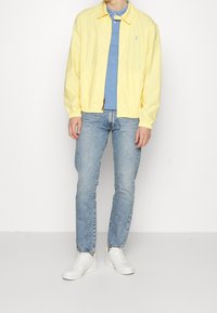 Hellgelbe, leichte Jacke mit Kragen, elastischen Bündchen und einem Reißverschluss auf der Vorderseite. Getragen über einem blauen Poloshirt, kombiniert mit hellblauen Jeans und weißen Turnschuhen.