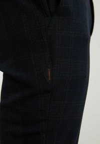 Marinblå rutiga byxor med en subtil textur, med en svart etikett med "CHASING" i orange på sidofickan.