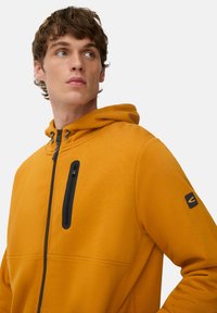 Sweat-shirt à capuche en tissu orange vif, avec une poche poitrine zippée noire et un patch logo sur la manche gauche. Texture lisse.