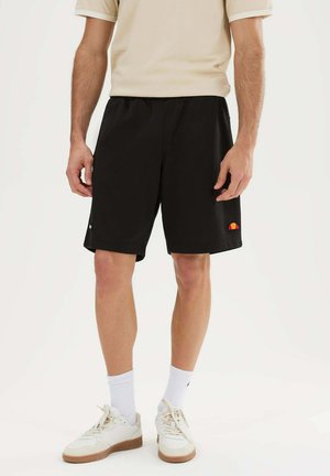 Mann trägt schwarze Shorts mit Logo, beiges Hemd, weiße Socken und weiße Turnschuhe und steht vor einem einfarbigen Hintergrund.