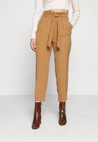Pantalon taille haute couleur sable avec ceinture à la taille, poches latérales et coupe fuselée, associé à des bottines marron texturées.