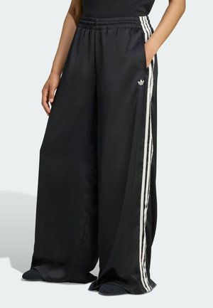 Osoba nosząca czarne szerokie spodnie Adidas z białymi paskami po bokach i elastycznym pasem, ręce w kieszeniach, stojąca na jasnym tle.
