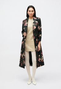 Pikk must lilleline mantel roosade ja rohelistena rõhutustega, kantud üle kuldse kootud kleidi, koos koorevärviliste sukkpükste ja teravate valgete kingadega.