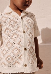 Virkatusta painonapillisesta paidasta, jossa on kerma- ja ruskeansävyinen timanttimalli, lyhyet hihat ja puiset napit. Pintakuvio on tarkasti yksityiskohtainen.