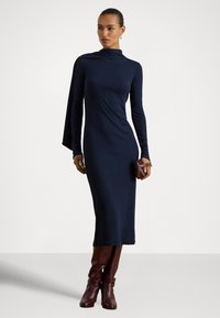 Ilga rankovė, aptempta tamsiai mėlyna suknelė su aukštu kaklu ir klostėta konstrukcija. Derinama su bordo spalvos kelius siekiančiais batais ir maža rankine. Lygi tekstūra.