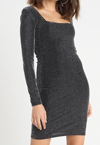 Zwart langshirt met een vierkante halslijn. Kenmerkt zich door een geribbelde textuur en zilveren verticale strepen voor een glinsterend effect. Bodycon pasvorm.
