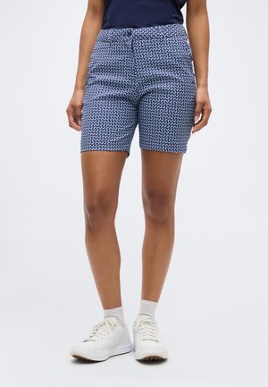 Persoană care poartă pantaloni scurți până la genunchi, cu model albastru închis și alb, șosete albe și adidași albi, stând în fața unui fundal simplu.