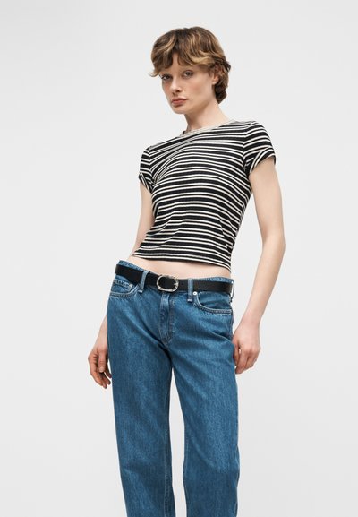 Νεαρό άτομο με κοντά, κυματιστά μαλλιά που φοράει μαύρο και λευκό ριγέ κοντό μπλουζάκι, μπλε τζιν και μαύρη ζώνη, στέκεται μπροστά σε λευκό φόντο.