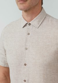 Kurzarm-Hemd mit Knopfleiste aus hellbeigem Stoff, das ein dezentes Streifenmuster und runde braune Knöpfe aufweist. Klassisches Kragen-Design.