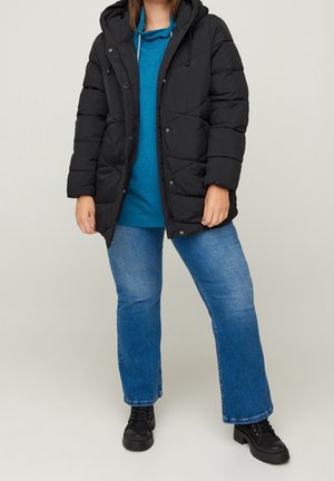 Personne portant une doudoune noire, un sweat à capuche bleu, un jean bleu et des bottes noires épaisses, debout devant un fond clair uni.