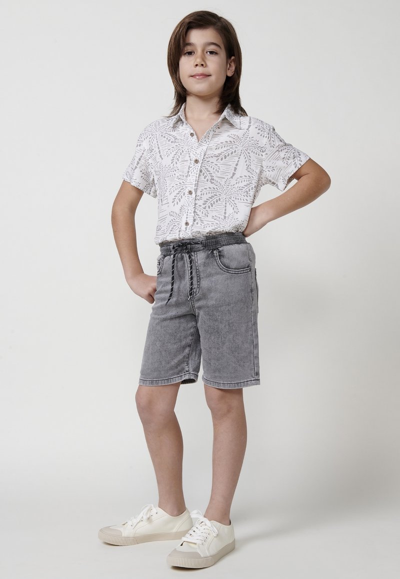 Camisa blanca de manga corta con estampado de hojas, botones claros; pantalones cortos de mezclilla gris con cinturilla elástica; zapatillas blancas con suelas beige.