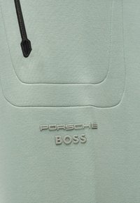 Υφασμα ανοιχτού πράσινου χρώματος με μαύρο φερμουάρ και ανάγλυφο κείμενο "PORSCHE BOSS" ραμμένο κάτω από το περίγραμμα της τσέπης.