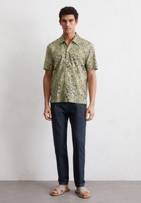 Korte mouwen, olijfgroene blouse met knopen en zwart bloemenpatroon, gecombineerd met donkerblauwe broek en grijze sandalen. Eenvoudig, casual ontwerp.