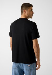 Zwart katoenen T-shirt met korte mouwen, ronde hals en een rechte pasvorm. Draagt een lichtblauwe spijkerbroek met achterzakken.