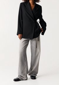 Veste blazer noire portefeuille à manches longues avec détail noué, associée à un jean gris clair à jambes larges. Chaussures noires à finition texturée.