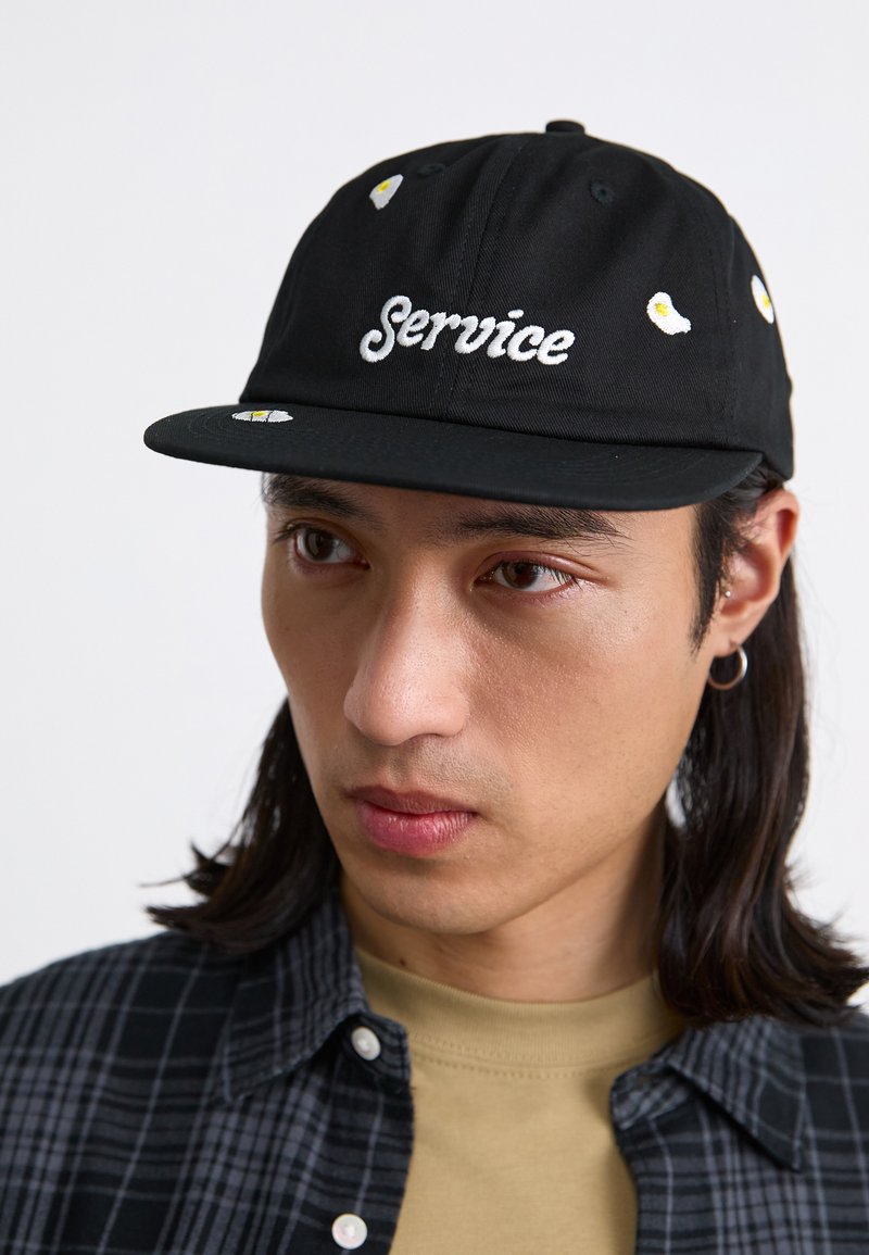Black cap with embroidered "Service" text and egg designs. Features a flat brim and button on top, made of soft material and structured shape.