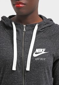 Sudadera gris con cremallera hecha de material suave, con un logo blanco y el texto "JUST DO IT" en el pecho, y cordones blancos en la capucha.