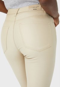 Ljust beige jeans med en slät yta, femficksdesign och synlig söm. Har en varumärkesetikett på midjebandet bak.