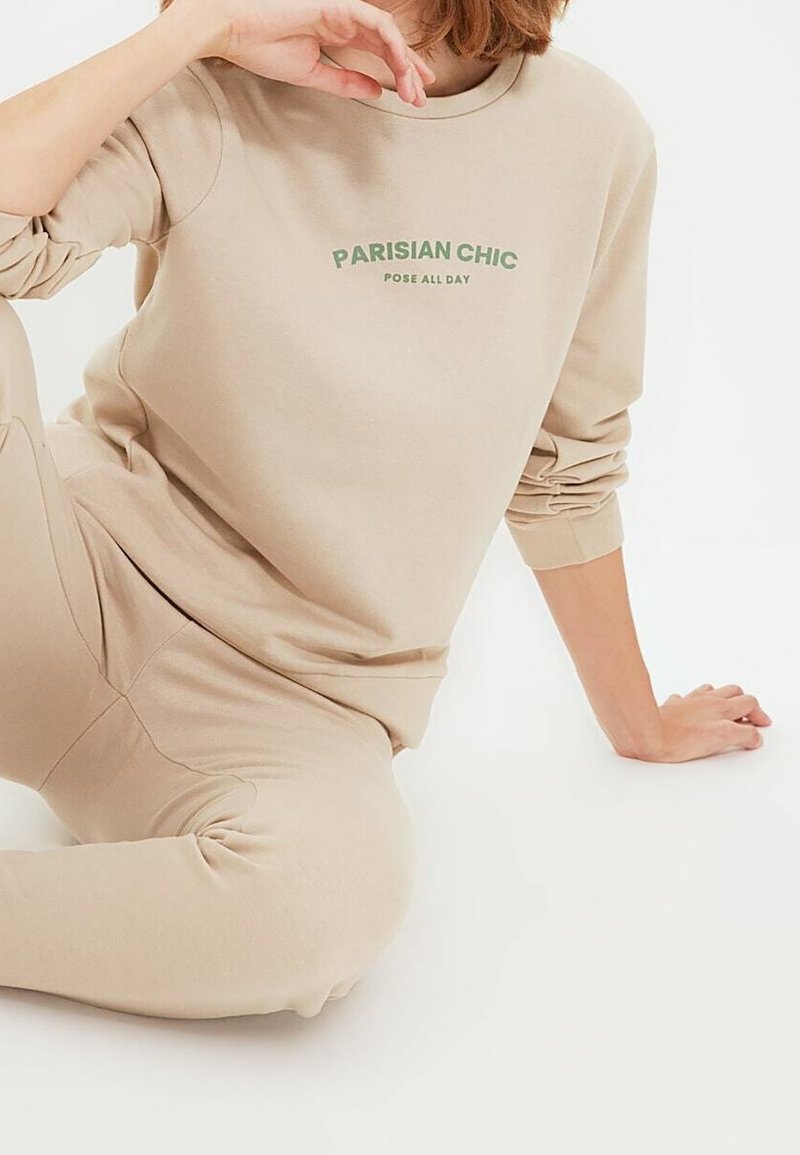 Persona seduta sul pavimento che indossa pantaloni e felpa beige con la scritta "PARISIAN CHIC POSE ALL DAY" stampata in lettere verdi.