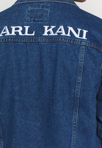 Džínová bunda ve středně modrém odstínu, s bílým vyšívaným textem "KARL KANI" na zadní straně. Klasický límec, zapínání na knoflíky a viditelné boční švy.