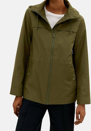 Personne portant une veste zippée vert olive avec une capuche et des poches à boutons-pression par-dessus une chemise blanche et un pantalon noir.