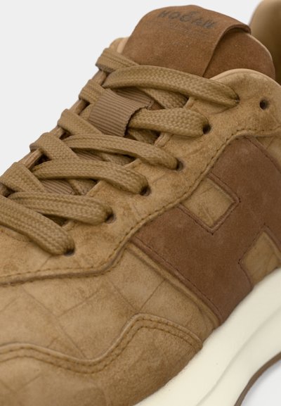 Brūnas zamšādas sporta apavi ar fakturētu augšdaļu, gludām ādas detaļām, plakanu šņori un polsterētu mēli. Mēlēs ir kontrastējoša logotipa izšuvums.