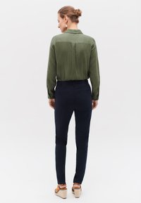 Grön långärmad skjorta med ryggskärning, kombinerad med marinkolumnära slim-fit byxor. Bruna kilklackar med vävd detalj. Neutral bakgrund.