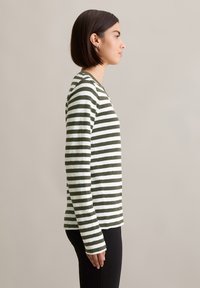 Woman with short brown hair wearing a long-sleeve green and white striped shirt, standing in profile against a plain beige background.