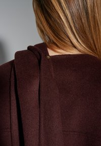 Burgundy wool blend garment featuring a draped design and a visible button closure at the shoulder, with a soft, textured finish.
