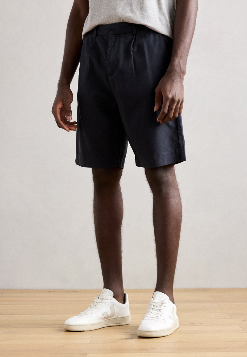Pantaloni scurți bleumarin cu o croială lejeră, cu pliuri în față, buzunare laterale și un design casual, asortați cu adidași albi pe un podea din lemn.