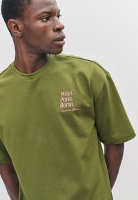 Olijfgroen katoenen T-shirt met korte mouwen, met roze tekst: "Milaan. Parijs. Berlijn. Euphoria Waves" op de linkerborst.