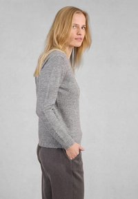 Donna con lunghi capelli biondi, che indossa un maglione grigio e pantaloni grigio scuro, in piedi di profilo con una mano in tasca contro uno sfondo semplice.