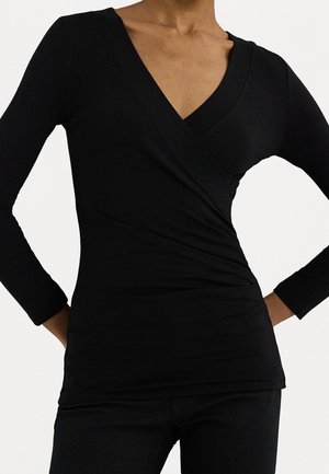 Vrouw draagt een nauwsluitende zwarte lange mouw wikkelshirt met een diepe V-hals, gecombineerd met een zwarte broek, handen rustend op de heupen.