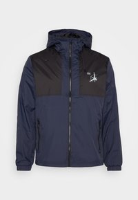 Giacca a vento blu navy con parte superiore a pannelli neri, chiusura con zip anteriore, due tasche laterali e cappuccio, con un piccolo logo sul petto.