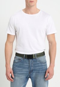 Balta kokvilnas krekliņš ar īsām piedurknēm, valkāts ar ziliem džinsu biksēm un zaļu jostu, uz kuras ir melns uzraksts "URBAN CLASSICS".