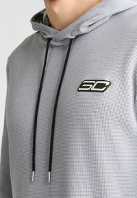 Hombre que lleva una sudadera con capucha texturizada de color gris claro, con cordones negros y un logo en blanco y negro "SC" en el pecho.