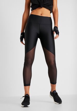 Vrouw draagt zwarte high-waist leggings met doorschijnende panelen, zwart cropped topje met knoop, vingervrije handschoenen en zwarte sportschoenen.