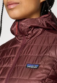 Gros plan sur une personne portant une veste matelassée Patagonia bordeaux avec zip et capuche, affichant le logo sur la poitrine.