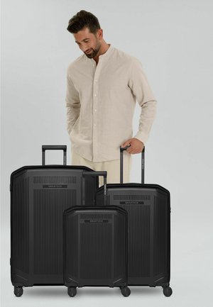 Mann in beigehemde steht mit drei schwarzen SMARTBOX-Koffern unterschiedlicher Größe mit ausgezogenen Griffen vor einem einfarbigen Hintergrund.