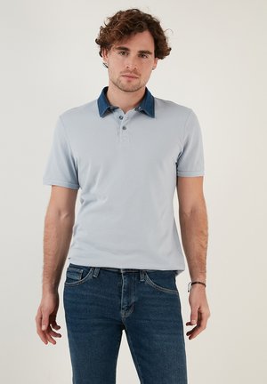 Vaaleansininen polo-paita, joka on valmistettu tekstuurilla varustetusta kankaasta, denim-kolarilla ja kolmella napilla varustetulla etuosalla. Yhdistetty tummansinisiin farkkuihin.