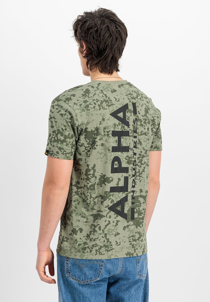Άνδρας που φοράει πράσινο t-shirt με μοτίβο παραλλαγής και μεγάλο κατακόρυφο κείμενο "ALPHA INDUSTRIES" στην πλάτη, συνδυασμένο με μπλε τζιν.