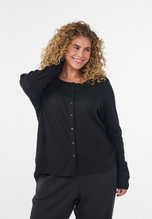 Mujer sonriente con cabello rubio rizado que lleva un top negro de manga larga con botones y pantalones negros, posando con una mano detrás de la cabeza.