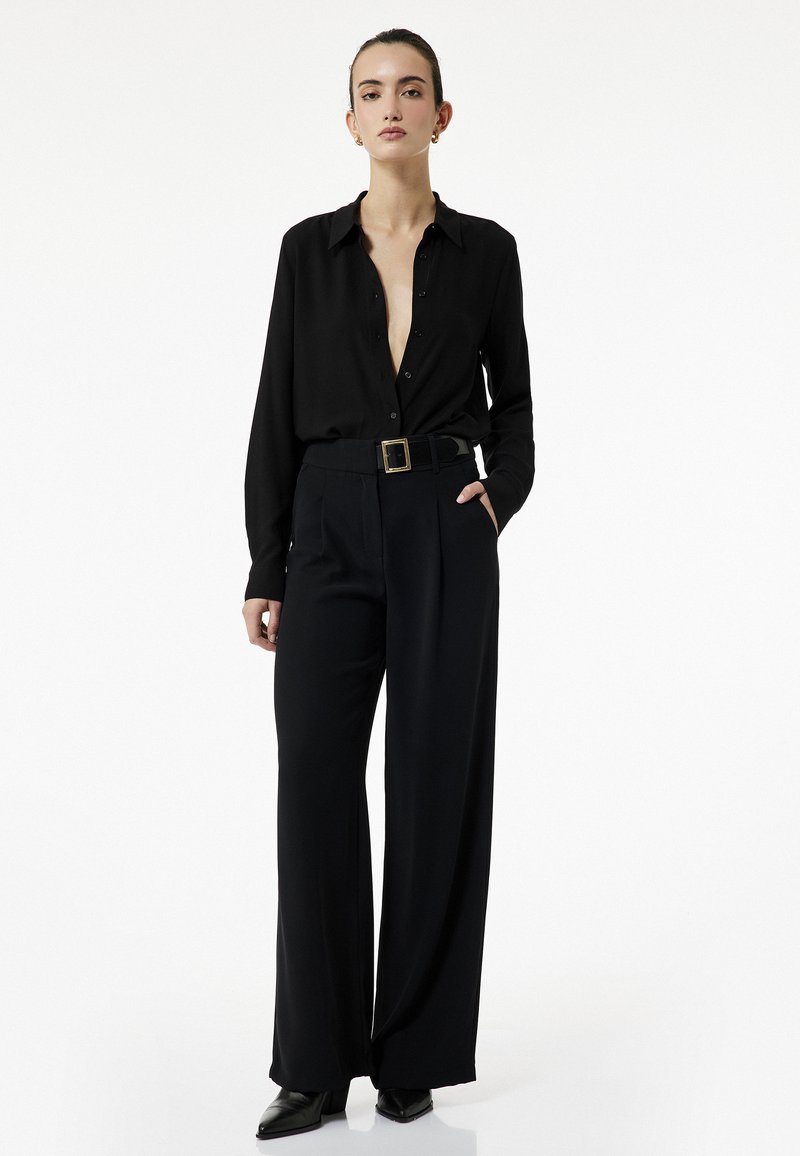 Schwarzes Langarmshirt mit tiefem V-Ausschnitt, kombiniert mit hoch taillierten schwarzen weiten Hosen und spitzen schwarzen Absätzen. Gürtel mit goldener Schnalle.