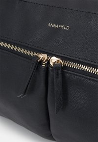 Černá kožená kabelka se zlatě tónovaným zipem a malými jezci. Má strukturovaný povrch a vyražené logo značky.
