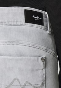 Szare ogrodniczki z dżinsu z czarną skórzaną metką marki, dwoma kieszeniami i ozdobnymi szwami, o fakturowanej tkaninie.