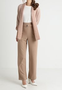 Jasnoróżowy prążkowany kardigan na białej koszuli, zestawiony z beżowymi spodniami z wysokim stanem, ozdobionymi paskiem i szerokimi nogawkami. Białe szpilki.