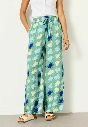 Woman wearing loose green pants with blue and cream sun patterns, a white sleeveless top, sandals, and bracelets on one wrist.