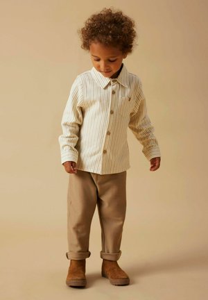 Gestreiftes Button-Up-Hemd in Creme mit Kragen, kombiniert mit hochgerollten braunen Hosen und braunen Stiefeln, vor einem neutralen Hintergrund.