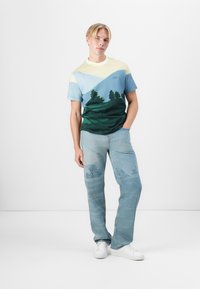 Grafická tričko se vzorem krajiny se zelenými stromy a modrými horami. Spárováno se světlými modrými džíny a bílými teniskami.