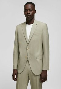 Veste de costume vert clair en tissu texturé avec un revers cranté, deux boutons et une poche poitrine, associée à une chemise blanche.